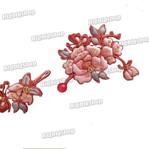 Pode incluir: Dois acessórios de cabelo florais decorativos. Um é um conjunto de flores rosa e brancas com detalhes dourados e folhas cinzentas. O outro é um design de borboleta e flor em tons de vermelho e rosa.