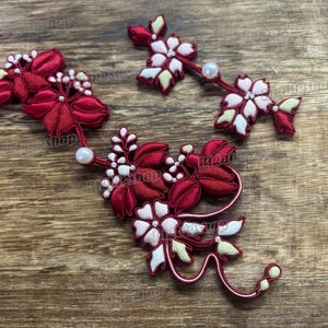 May include: Three red and white floral hair accessories with pearl accents are arranged on a wooden surface. The flowers have a layered petal design and are connected by thin, red stems. The accessories are likely handmade.