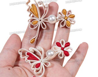 Op de afbeelding: Twee paar decoratieve stoffen versieringen in de vorm van vlinders en bloemen. De vlinders zijn wit met rode en gouden accenten en de bloemen zijn wit met rode accenten. Elke versiering heeft een parel in het midden.