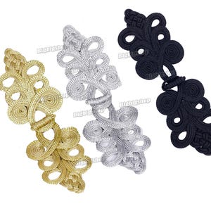 May include: Three decorative frog closures in gold, silver, and black. Each closure features an intricate, looped design, suitable for embellishing garments or accessories. The closures are made of a woven material.