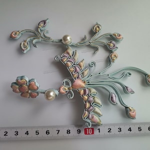 May include: Two embroidered floral appliques with a bird-like design. The appliques feature light blue stems, pink, gold, and purple accents, and faux pearls. A ruler shows the appliques are approximately 4 inches wide.