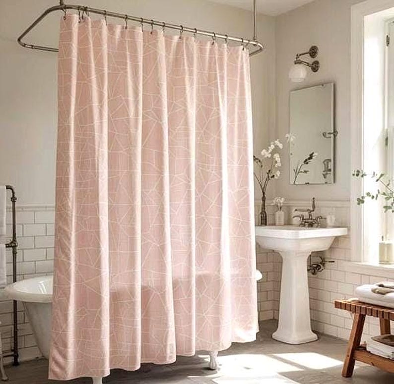 May include: A pink shower curtain with a white geometric pattern hangs in a bathroom. The curtain is suspended from a curved metal rod. The bathroom has a white sink, mirror, and a wooden bench.