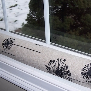 May include: A beige fabric draft stopper with a black dandelion pattern rests on a white windowsill. The cylindrical design is positioned in front of a window with a snowy outdoor view.