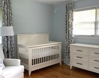 May include: A white cot with a white mattress and a white chest of drawers with six drawers in a light blue nursery with a white rocking chair and white curtains with a black and white pattern.