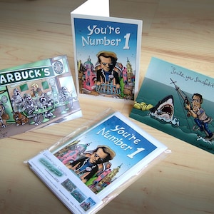 May include: A collection of Geekings Cards greeting cards featuring humorous illustrations. One card reads "You're Number 1", another depicts a scene at Starbucks with Star Wars characters, and a third shows a man on a water ski being chased by a shark.