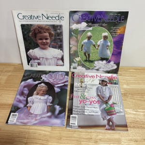 May include: Four vintage issues of "Creative Needle" magazine are displayed. The covers feature children in white dresses and bonnets, with floral and pastel backgrounds. The magazine covers have the text "Creative Needle" and various issue titles.