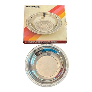 Könnte beinhalten: Zwei silberfarbene Servierschalen mit dekorativen Rändern und geprägten floralen Mustern. Eine Schale befindet sich in der Originalverpackung, auf der der Markenname "IRVINWARE" aufgedruckt ist. Die Box hat ein Retro-Farbschema aus Rot, Orange und Gelb.