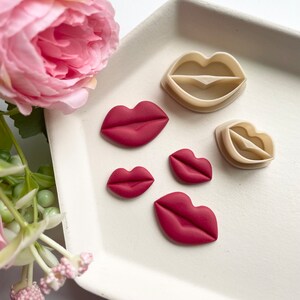 Peut inclure: Quatre breloques en argile polymère en forme de lèvres rouges et deux emporte-pièces en plastique en forme de lèvres sur une assiette blanche. Les breloques sont de tailles différentes.