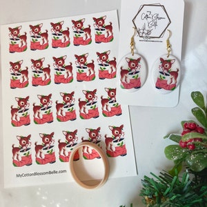 May include: A sheet of stickers featuring a vintage illustration of a reindeer in a red boot holding candy canes. The stickers are arranged in a grid pattern. There are also two white ceramic earrings with the same reindeer illustration. The earrings are attached to gold hooks. The text "MyCottonBlossomBelle.com" is printed on the sheet of stickers.
