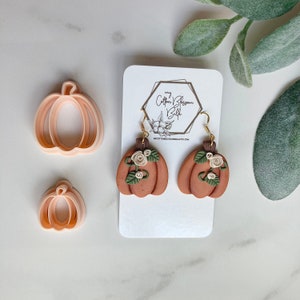 May include: A pair of brown pumpkin earrings with white flowers and green leaves. The earrings are hanging from a white card with the text "my Cotton Blossom Belle" and a website address.