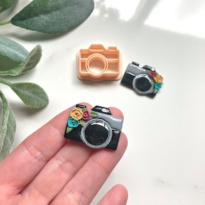 May include: A black and grey camera-shaped charm with colourful floral accents. The charm has a large lens and a flash. A matching beige-coloured camera-shaped cookie cutter is also visible. The charm is approximately 2.5 cm in length.