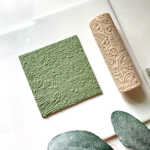 May include: A beige rolling pin with a mushroom and leaf design. The rolling pin is used to create an embossed pattern on a green square of clay.
