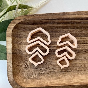 May include: Two pink plastic cookie cutters shaped like a stylized arrow design. The cutters are on a wooden surface.