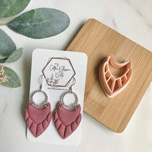 May include: A pair of mauve polymer clay earrings with a silver hoop and a scalloped design. The earrings are displayed on a white card with the text "my Cotton Blossom Belle" and a website address. A pink polymer clay cookie cutter in the shape of a crescent moon is visible in the background.