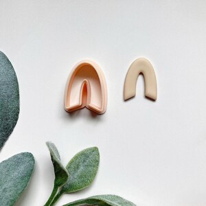 May include: A light pink plastic cookie cutter shaped like an arch, and a light brown clay arch. The cookie cutter is slightly larger than the clay arch.