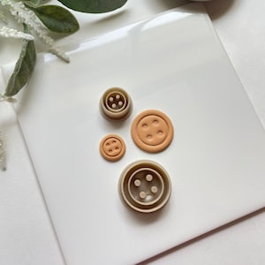 Peut inclure: Quatre emportes-pièces en plastique de différentes tailles en forme de boutons. Les emportes-pièces sont beiges et présentent un motif en relief de quatre trous au centre. Les emportes-pièces sont sur une surface blanche.