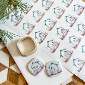 May include: Two square ceramic ornaments with a white background and a watercolor illustration of the nativity scene. The ornaments have a gold glitter border and are on a white surface.