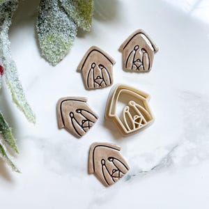 May include: A collection of beige house-shaped cookie cutters with a nativity scene design. The cutters are arranged on a white surface, with a sprig of green foliage in the background. The cutters are made of plastic.
