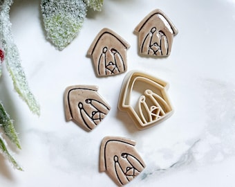 May include: A collection of beige house-shaped cookie cutters with a nativity scene design. The cutters are arranged on a white surface, with a sprig of green foliage in the background. The cutters are made of plastic.