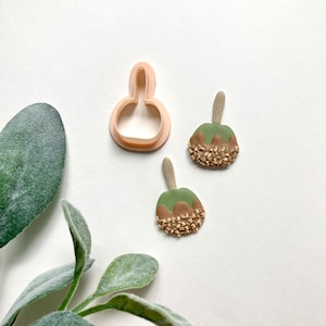 May include: Two green and brown candy apple shaped polymer clay earrings with a brown stick on top. A pink plastic cookie cutter in the shape of an apple is also in the image.