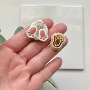 May include: A set of three white polymer clay bunny foot cookie cutters. The cutters are in the shape of bunny feet with pink paw pads. The cutters are on a white surface.