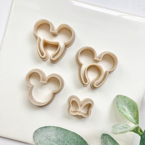 May include: Four plastic cookie cutters in the shape of a bow, a mouse head, and two different styles of mouse ears. The cutters are beige and are on a white surface.