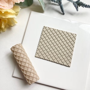 May include: A beige rolling pin with a diamond pattern and a beige square tile with a diamond pattern. The tile and rolling pin are on a white surface.