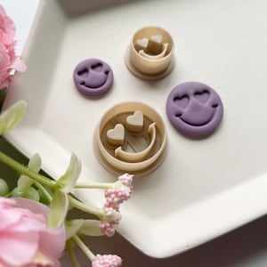 May include: Three cookie cutters in the shape of a smiley face with hearts for eyes. Two of the cutters are purple and one is beige.