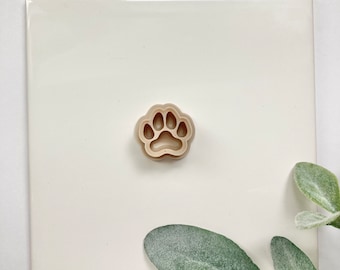 May include: A beige plastic paw print cookie cutter. The cutter is on a white surface.