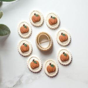 May include: A collection of fall-themed craft supplies. The image features eight oval-shaped, off-white decorations with orange pumpkin designs and green leaves. A pumpkin-shaped cookie cutter is in the center. The items are arranged on a white surface.