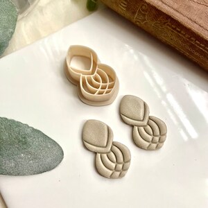 May include: A set of two beige clay earrings with a geometric design. The earrings are made with a mold and have a textured surface. The earrings are shown next to a plastic mold.
