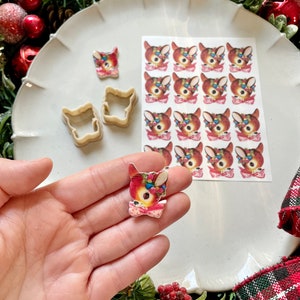 May include: A sheet of edible images featuring a vintage-style cartoon deer with a red bow and a wreath of holly and berries. The images are printed on a white background and are ready to be used for decorating cakes, cupcakes, or other desserts. There are two cookie cutters in the shape of a deer head, and a single deer image is being held in a hand.