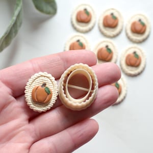 May include: A pumpkin-shaped cookie cutter and a decorated cookie with an orange pumpkin design. The cookie has a beige background with a scalloped edge. Several more cookies with pumpkin designs are in the background.