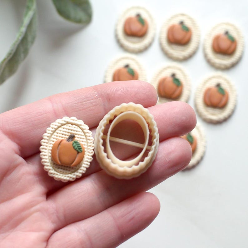 May include: A pumpkin-shaped cookie cutter and a decorated cookie with an orange pumpkin design. The cookie has a beige background with a scalloped edge. Several more cookies with pumpkin designs are in the background.