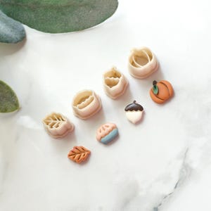 May include: A selection of autumn-themed cookie cutters in shapes such as a leaf, acorn, pumpkin, and pie. The cutters are beige and brown, with green and blue details. Perfect for making seasonal biscuits and treats.