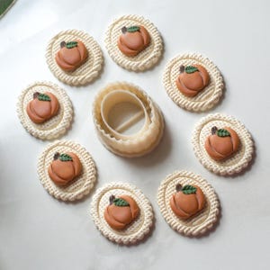 May include: A collection of pumpkin-shaped cookies with green leaf details, arranged around a beige oval cookie cutter. The cookies are set on a white surface, showcasing a fall theme. The cookies are approximately 2 inches in diameter.