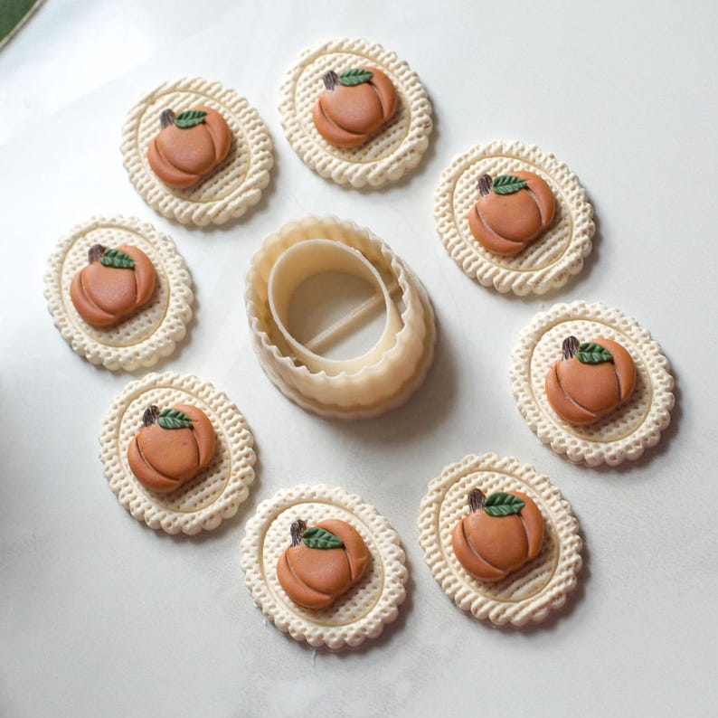 May include: A collection of pumpkin-shaped cookies with green leaf details, arranged around a beige oval cookie cutter. The cookies are set on a white surface, showcasing a fall theme. The cookies are approximately 2 inches in diameter.