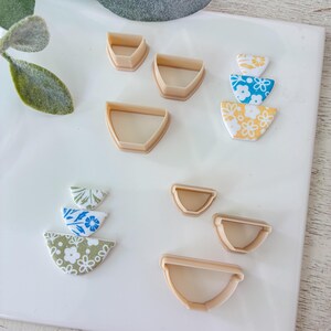 May include: A collection of beige plastic semi-circle clay cutters in various sizes alongside two stacked sets of semi-circular clay pieces featuring white floral patterns on yellow, blue, and green backgrounds arranged on a white surface.