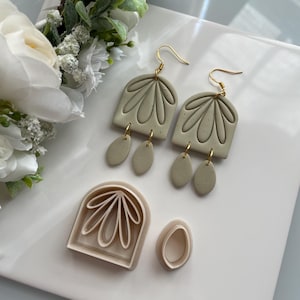 May include: A pair of sage green dangle earrings with gold-colored hooks and accents. The earrings feature an arched top with a floral design and three dangling oval shapes. Also included are matching clay cutters for creating the earrings.