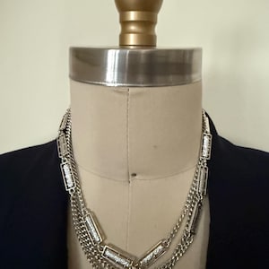 May include: A multi-strand silver-toned necklace featuring a combination of delicate curb chains and rectangular metal links with a textured, etched surface. The necklace is displayed on a beige mannequin torso wearing a dark navy blue blazer.
