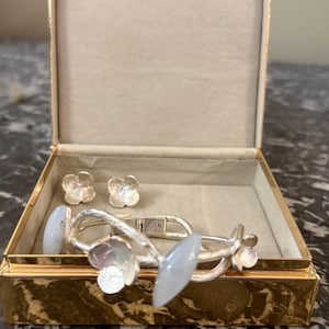May include: A gold jewelry box with a beige interior holds a silver bracelet and matching earrings. The bracelet features light blue oval and flower-shaped accents. The earrings are small flower studs. The box is open, revealing the jewelry inside.
