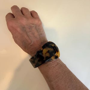 May include: A wide cuff bracelet featuring a classic tortoiseshell pattern with mottled brown, black, and amber tones. The bracelet has a smooth, polished finish and a curved, buckle-style design that wraps around the wrist with a solid, continuous band.