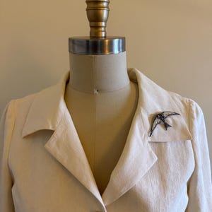 May include: A cream-colored blazer with a subtle tonal pattern, featuring a notched lapel and a single button closure. A silver-toned swallow bird brooch with black and white enamel detailing is pinned to the left lapel of the garment.