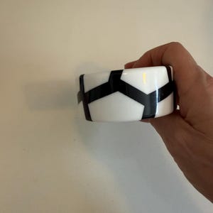 May include: A wide bangle bracelet featuring a high-contrast geometric pattern of solid black shapes set against a smooth white background. The design consists of interconnected angular forms wrapping around the circumference of the circular band.
