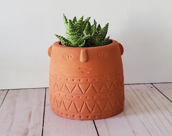 May include: A terracotta planter with a face design and geometric patterns. The planter is filled with a green succulent plant.