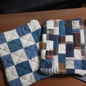 May include: Two quilted laptop sleeves and a pair of eyeglasses rest on a wooden table. One sleeve features a blue and white geometric pattern, while the other is a patchwork of various fabrics in blue, brown, and white. A silver laptop is also present.