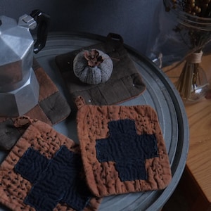 May include: A silver coffee pot sits on a brown and blue quilted pot holder. Two more brown and blue quilted pot holders are on a gray metal surface. A small brown and gray fabric ball sits on a brown and gray quilted pot holder.