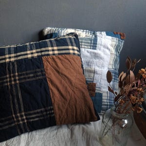 May include: Two square throw pillows with patchwork designs. One pillow features a navy blue and tan plaid pattern with a brown center panel. The other pillow has a blue and white patchwork design. Decorative home decor.