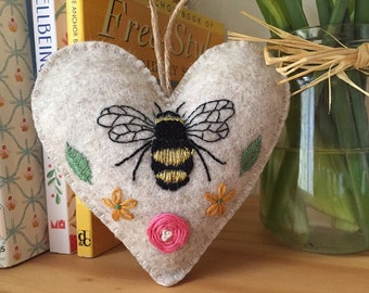 May include: A heart-shaped felt ornament in light beige featuring a hand-embroidered bumblebee in black and gold thread. The design includes two small green leaves, two orange flowers, and a central pink circular stitched accent with a twine loop.