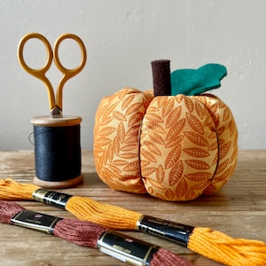 May include: A fabric pumpkin pincushion with a leafy pattern in shades of orange and brown. The pumpkin has a green felt stem and is sitting on a wooden surface. There are also spools of thread and a pair of scissors in the image.